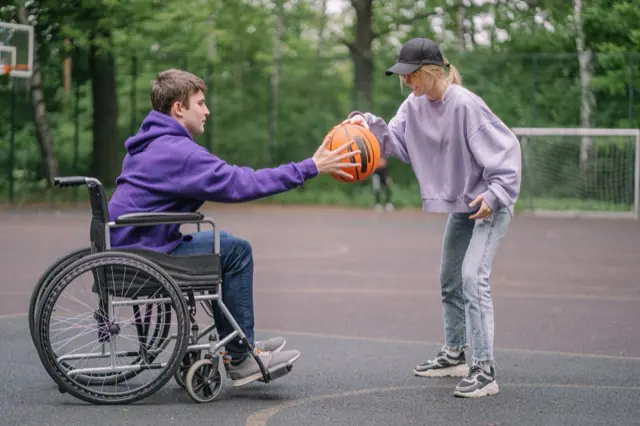 Two people playing basketball on an outdoor court, one of them using a wheelchair.