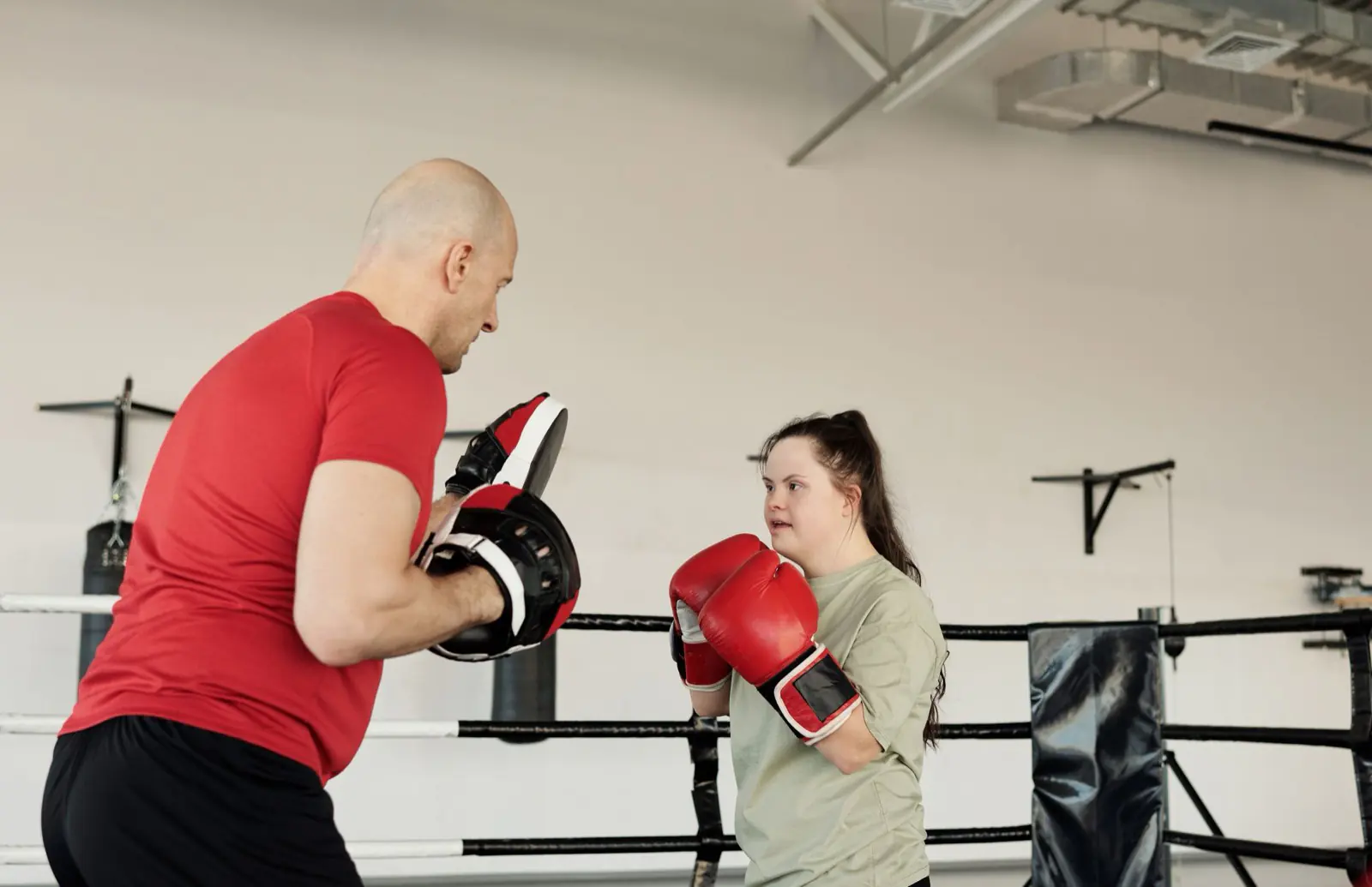 A trainer guiding a young woman with Down syndrome through a boxing session in a gym.