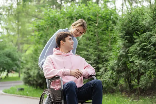 Two people on a garden path, one using a wheelchair, both smiling.