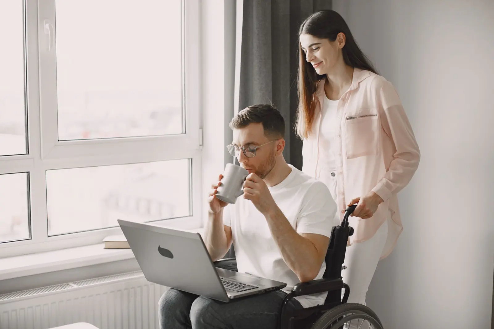 A man using a wheelchair working at a laptop with a coffee, a support worker nearby beside a bright window.