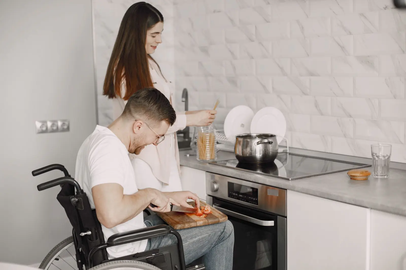 A man using a wheelchair preparing food at a kitchen bench alongside a support worker.