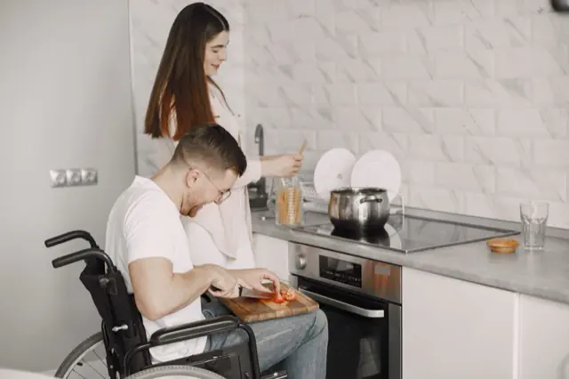 A man using a wheelchair preparing food at a kitchen bench alongside a support worker.