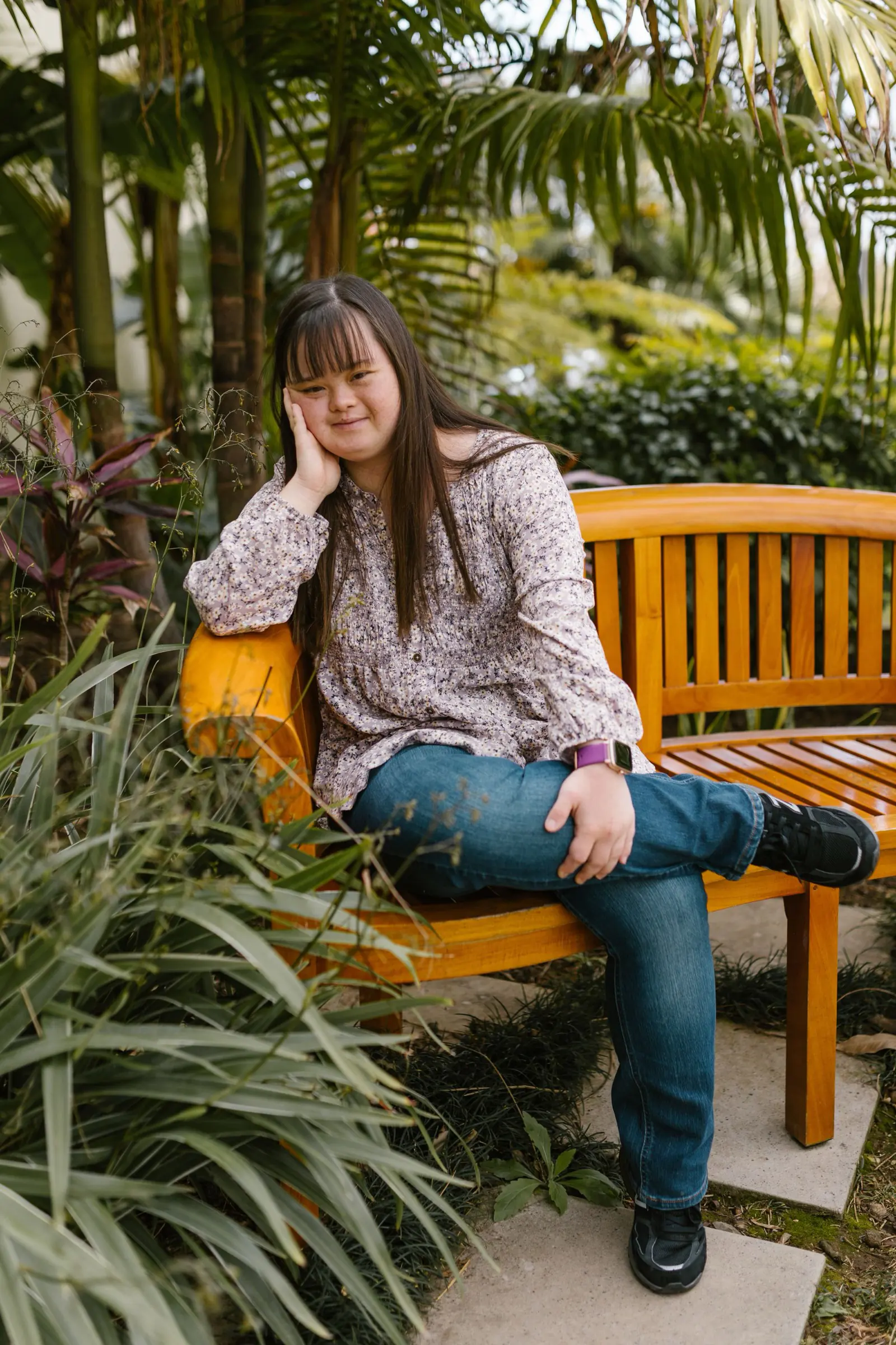 A young woman with Down syndrome resting on a yellow bench in a lush green garden.